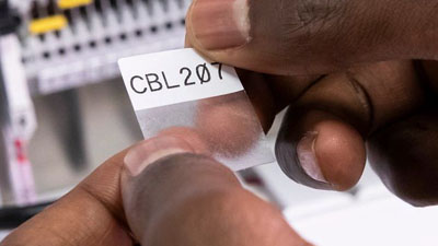 Een technicus brengt een zelflaminerend vinyllabel van Brady met extra sterke kleefstof aan op een draad of kabel. De tekst is duidelijk geprint op het witte labelgedeelte en er is een transparant gedeelte dat de tekst bedekt wanneer het wordt aangebracht.